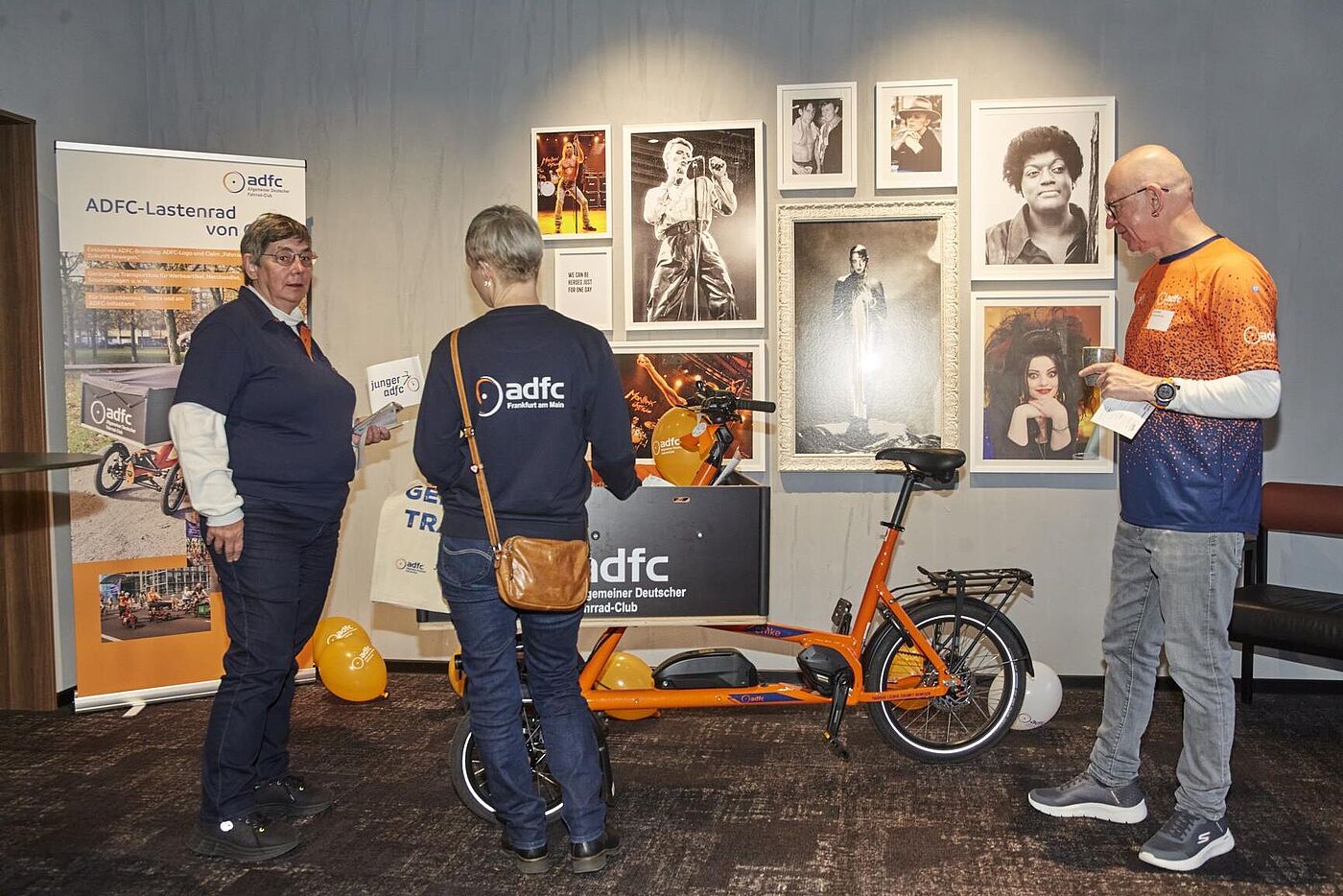 Drei Personen informieren sich über das orangefarbene ADFC‑Lastenrad. Im Hintergrund sind Roll‑ups zum ADFC‑Lastenrad, Luftballons und mehrere gerahmte Fotos von Musikerinnen und Musikern an der Wand zu sehen.