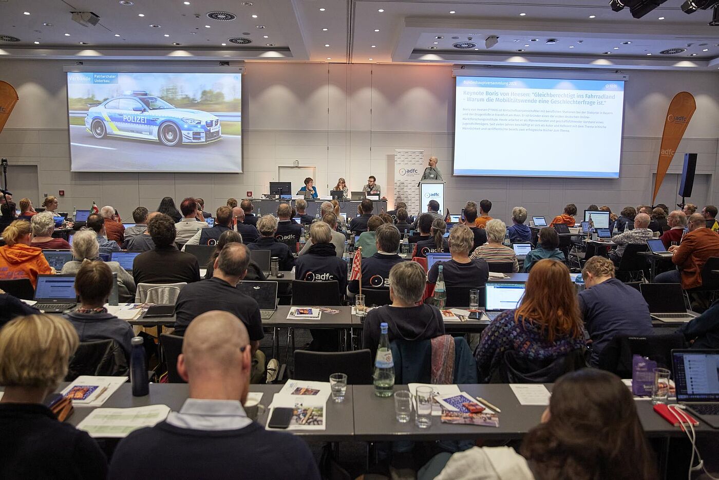 Teilnehmende sitzen mit Laptops im Saal und hören der Keynote von Boris von Heesen „Gleichberechtigt ins Fahrradland – Warum die Mobilitätswende eine Geschlechterfrage ist“ auf der Bühne mit zwei Projektionsleinwänden zu.