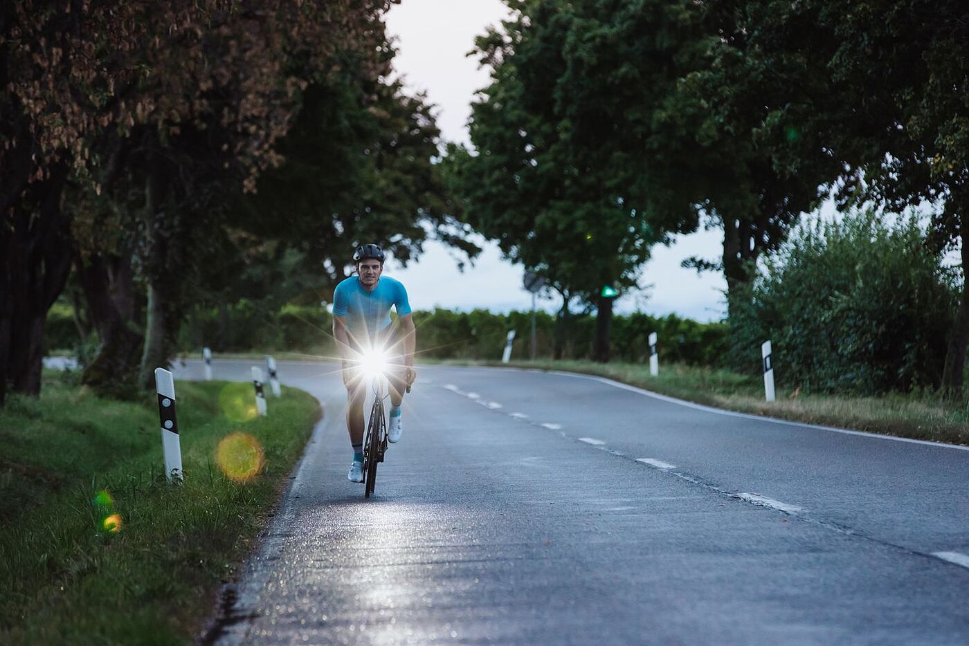 Ein Rennradfahrer fährt in der Dämmerung auf einer Landstraße auf die Kamera zu, am Lenker ein Scheinwerfer. Im Hintergund grüne Landschaft. 