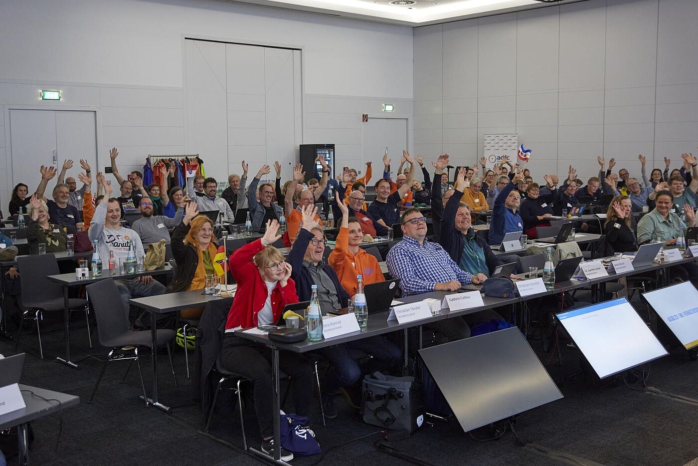 Die Teilnehmenden sitzen im Saal und heben gemeinsam Hände in die Luft.